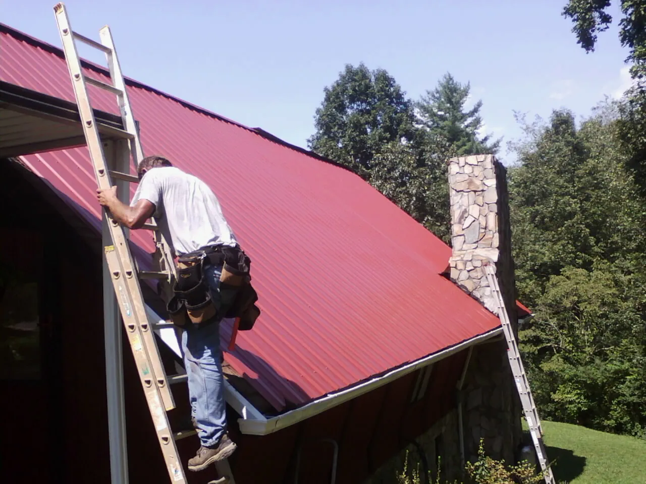 Roofer on a ladder beside a red metal roof and stone chimney — Soffit & Fascia Repair by Roofing Brothers in Lenore